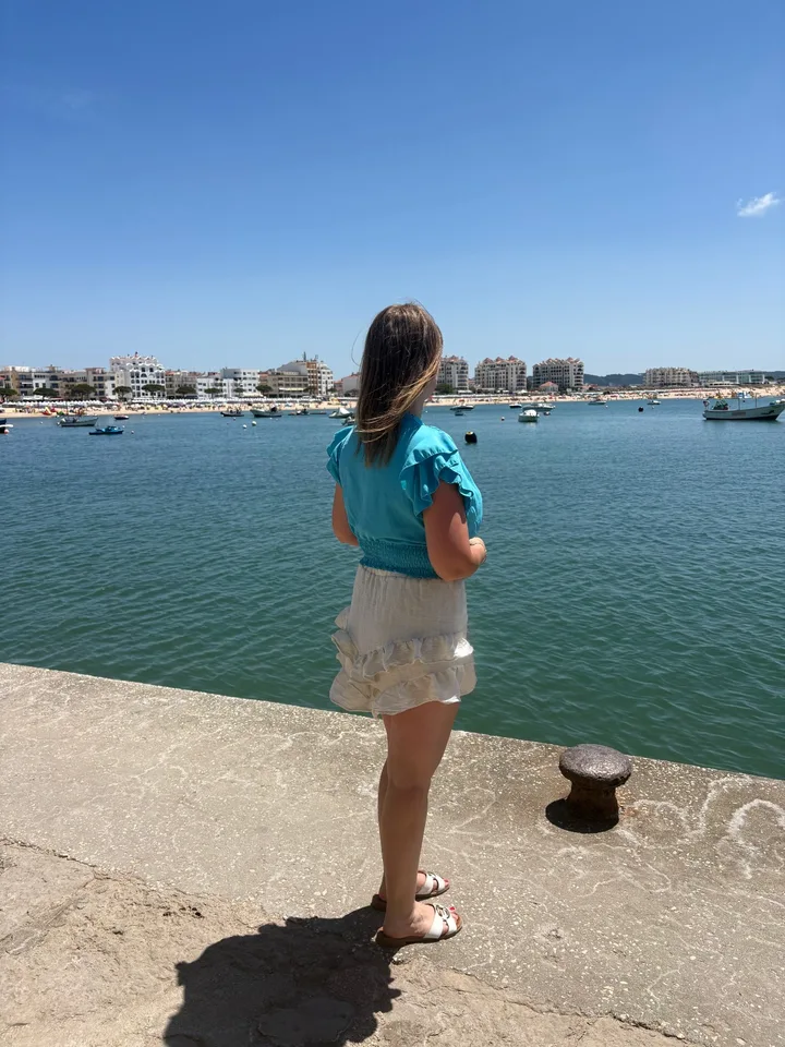 Demi kijkt uit over een haven, met blauwe lucht en boten op de achtergrond